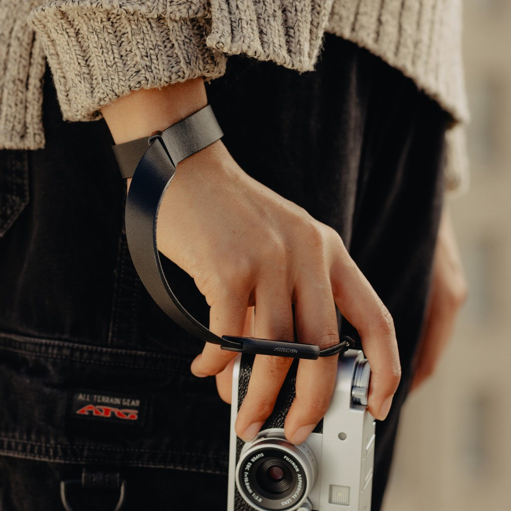 Person holding a camera with a Black Leather Cuff, wearing a textured sweater and dark pants. Person holding a camera with a Black Leather Cuff, wearing a textured sweater and dark pants.