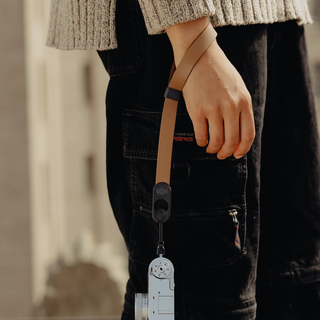 Person holding a camera with a Tan Leather Cuff, wearing black pants and a beige sweater. Person holding a camera with a Tan Leather Cuff, wearing black pants and a beige sweater.